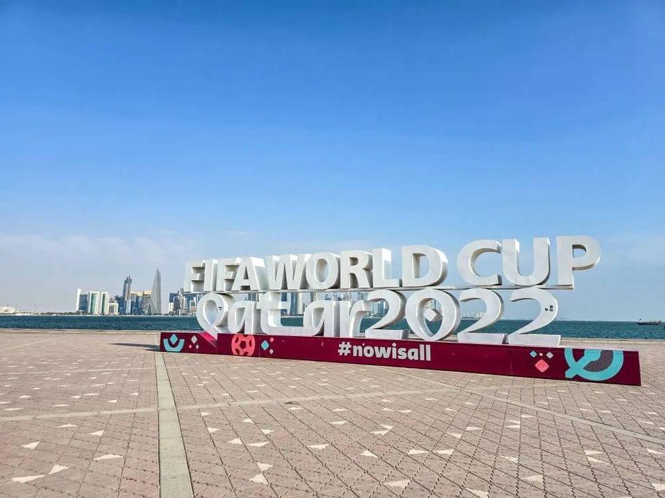 A Qatar 2022 sign stands beside blue water, with Doha’s towers across the bay.