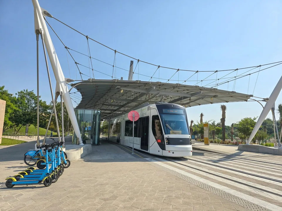 A white tram waits beneath a angular station canopy beside parked bicycles and scooters.