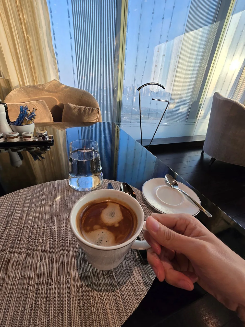 A hand holds a white cup of coffee in front of a bright window.