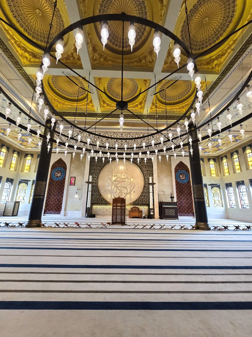 Circular hanging lamps illuminate a mosque interior with a decorated gold ceiling and patterned carpet.