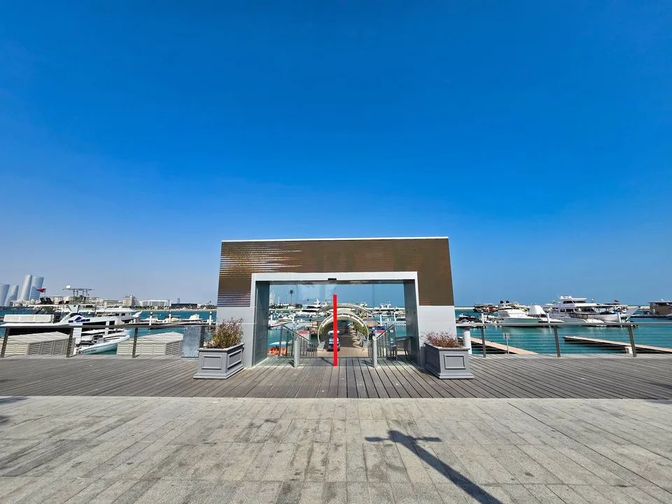 A rectangular metal entrance frame overlooks yachts and pale waterfront buildings at the marina.