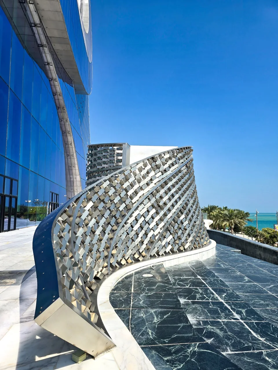 A curved woven-metal sculpture stands beside blue glass and a narrow water feature.