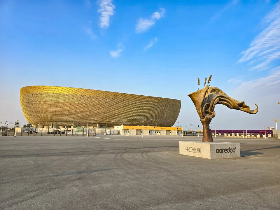 A gold-coloured stadium stands behind a large bronze sculpture on a paved plaza.