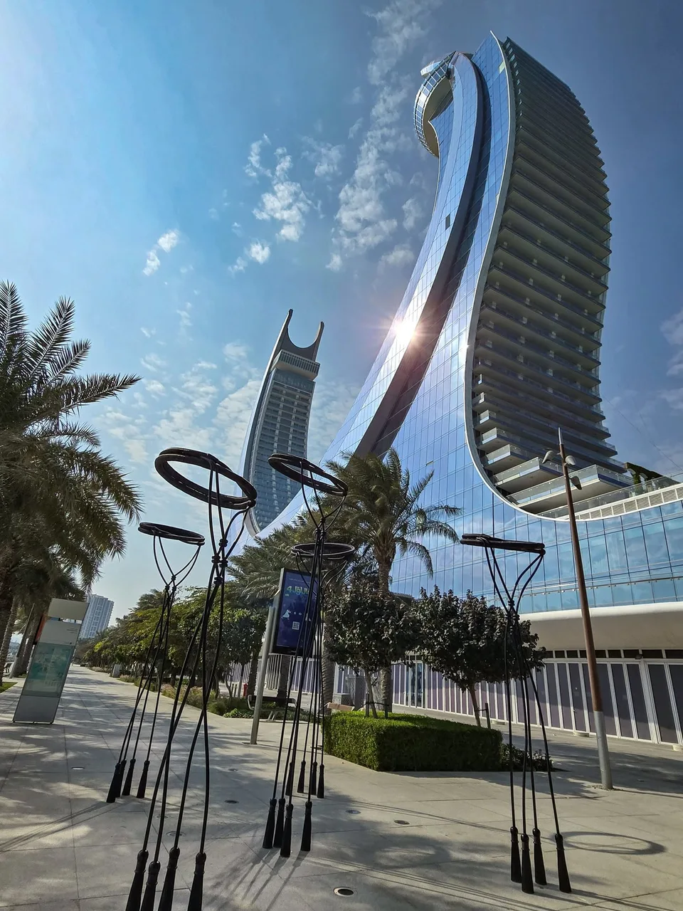 Slender dark sculptures and palm trees line an empty pavement beneath a curved glass tower.