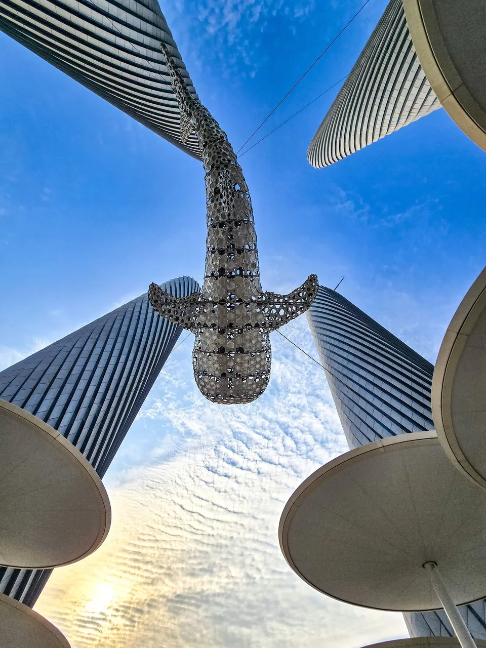 A suspended metal whale sculpture hangs overhead between four curved towers in Lusail.