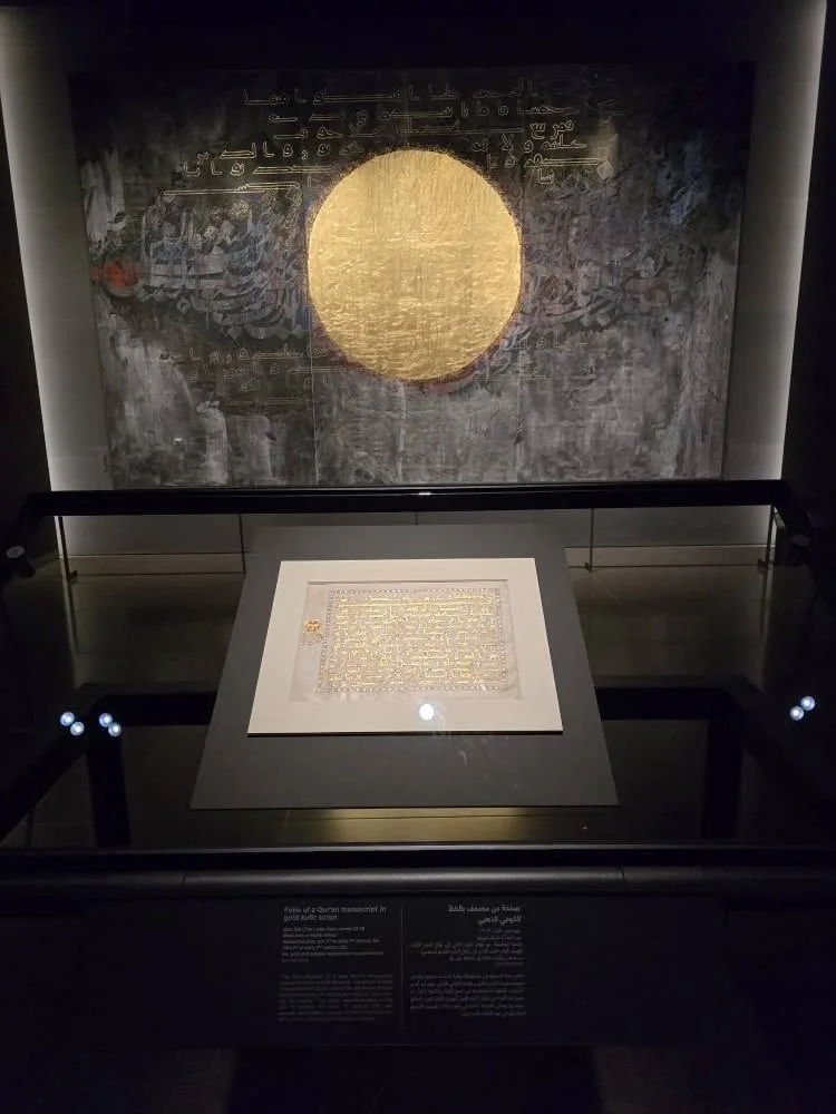 A circular gold calligraphic artwork hangs above a manuscript display in a dark gallery.