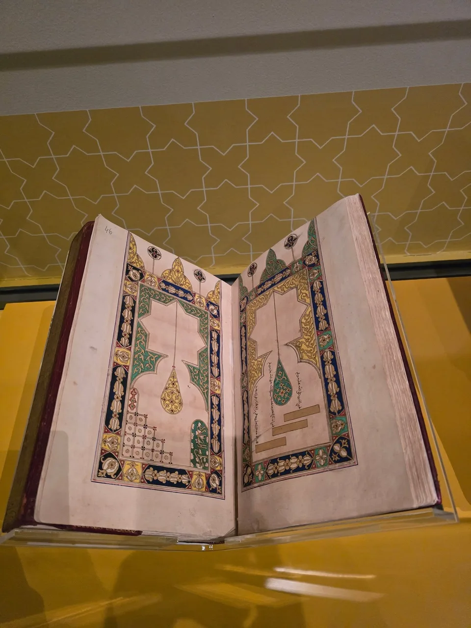 An open book displays small illustrations and Arabic text within intricate green, blue and gold borders.