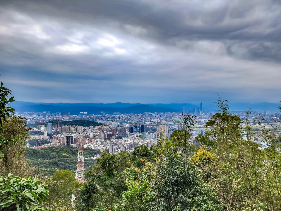 Taipei spreads beyond green slopes, with Taipei 101 in the distance under cloudy skies.