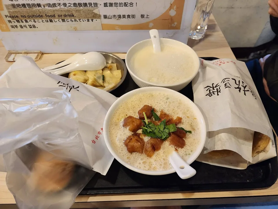 A bowl topped with youtiao pieces and green herbs sits on a tray beside another bowl and wrapped food.