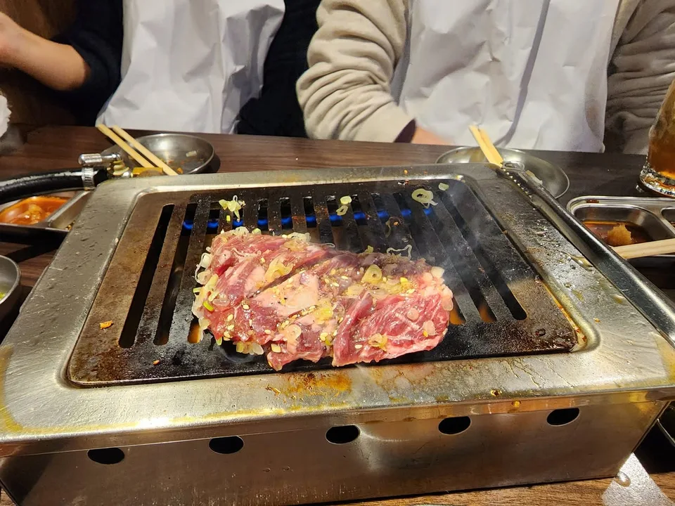 Marbled slices of meat topped with spring onions cook on a tabletop grill above blue flames.