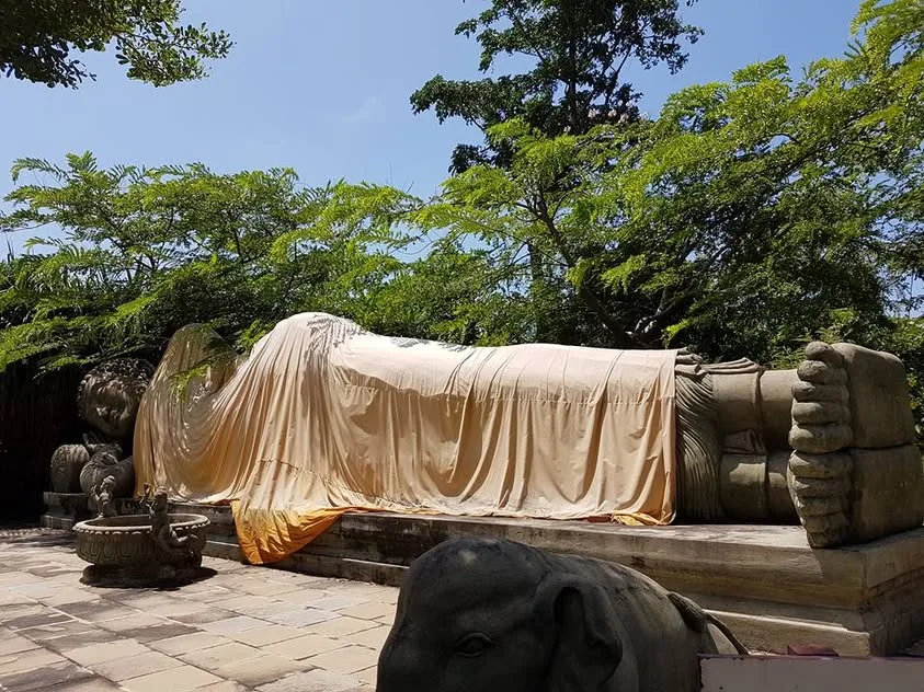 A reclining Buddha sculpture lies outdoors beneath trees, draped in bright cloth.