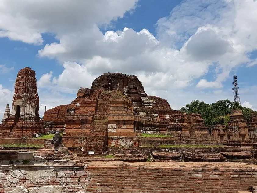 Weathered brick walls and temple structures stand beneath a bright sky, with a dark stone Buddha figure in the ruins.