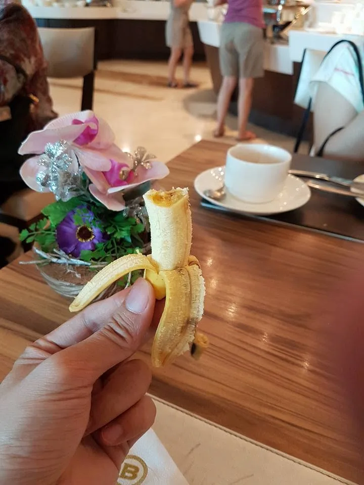 A hand holds a peeled banana above a wooden table beside a cup; other diners’ bodies are visible without their faces.