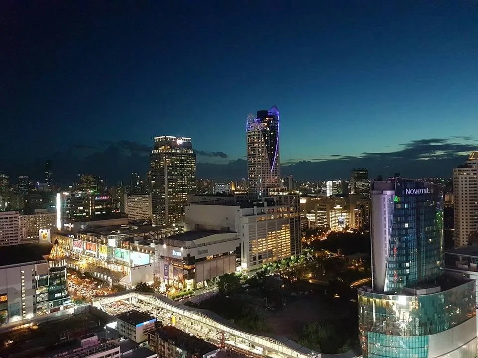 Tall illuminated buildings rise above Bangkok’s streets and skywalks, with CentralWorld’s name visible on a lower building.