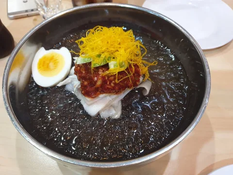 A metal bowl of cold noodles is topped with half a boiled egg, cucumber and a fine yellow garnish.