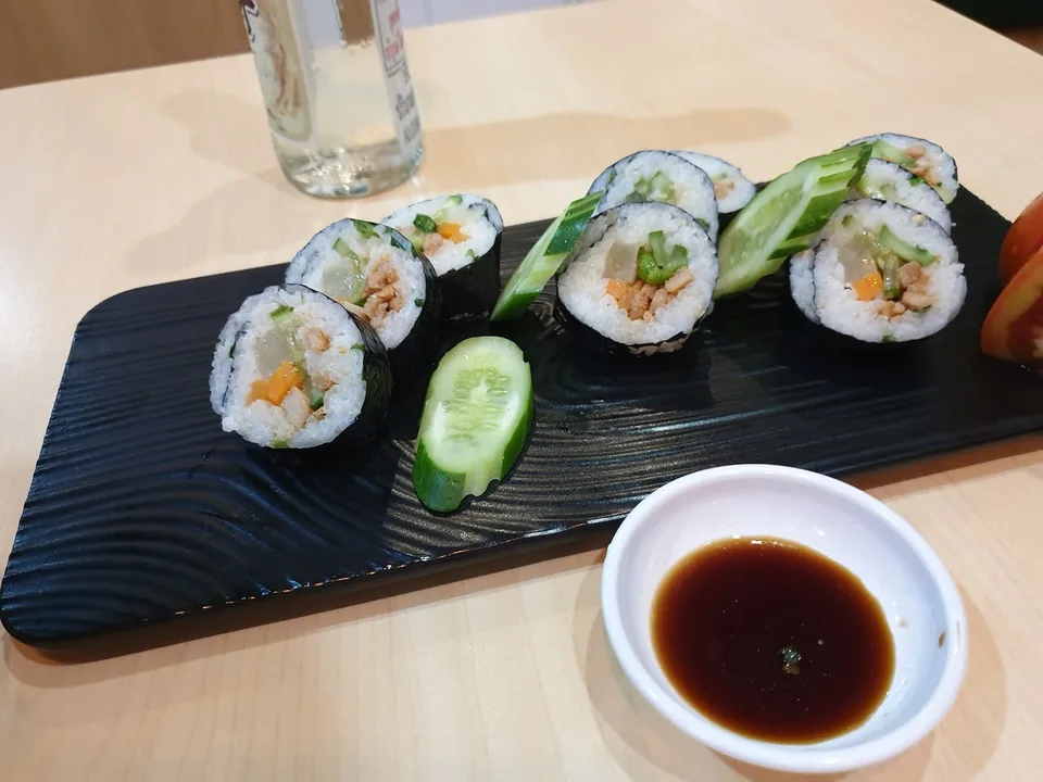 Sliced rice rolls and cucumber sit on a black platter beside a small bowl of dipping sauce.